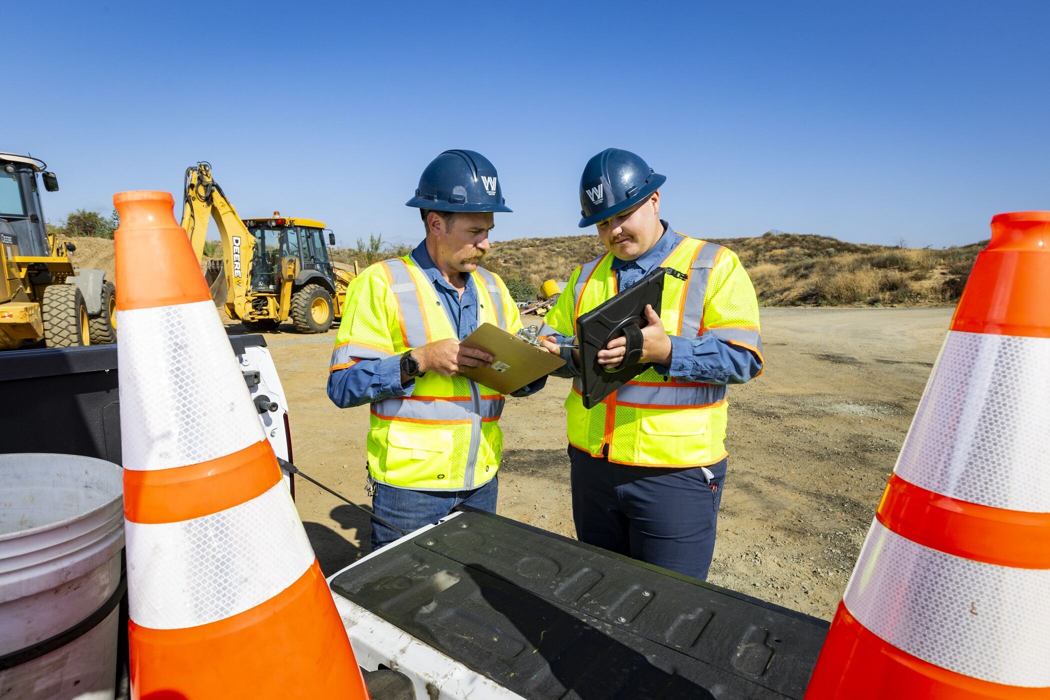 Two construction workers in blue hard hats and yellow vests discuss paperwork at a worksite.